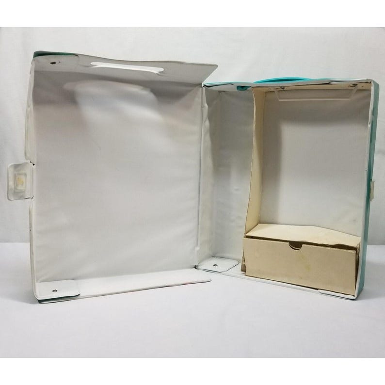 Op de afbeelding: Open vintage koffer met een wit interieur en een klein, beige compartiment. De buitenkant is lichtblauwgroen met een witte handgreep en een doorzichtige plastic sluiting. De koffer is open en onthult de binnenkant.