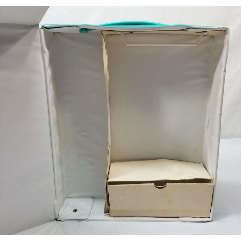 Op de afbeelding: Een open doos in vintage-stijl met een turquoise handvat en afwerking. De binnenkant is verdeeld in compartimenten, met een kleine, beige lade aan de onderkant. De doos is wit met een licht getextureerd oppervlak.