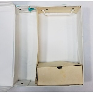 Op de afbeelding: Een open, witte vintage koffer met een kleine, beige kartonnen doos erin. De koffer heeft een wit interieur en een lichtblauwe rand. De doos heeft een cirkelvormige uitsparing aan de voorkant.