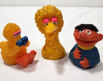 May include: Three small molded plastic figures featuring a yellow bird character holding a blue camera, a yellow bird character with pink eyelids, and an orange character with black hair wearing a blue sweater with a yellow collar.