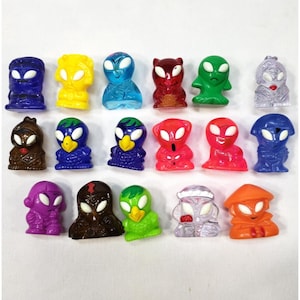 May include: A collection of colorful alien-like figurines in various colors, including blue, yellow, red, green, and purple. Each figurine has a unique design, with large eyes and distinct features. The figurines are made of a translucent material.