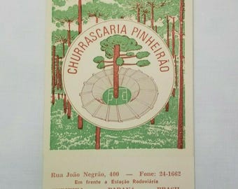 May include: A rectangular vintage postcard featuring a green forest illustration with a central circular emblem. The emblem depicts a stadium surrounding a large pine tree, with the text CHURRASCARIA PINHEIRÃO. Below, text lists an address and phone number in Curitiba, Parana, Brasil.