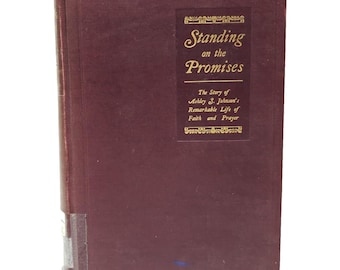 May include: A maroon hardcover book titled "Standing on the Promises." The book's cover features gold lettering and a decorative border. The text below the title reads "The Story of Ashley S. Johnson's Remarkable Life of Faith and Prayer."