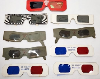 May include: A collection of nine paper 3D glasses arranged in rows. The set includes a red Coca-Cola branded pair, a black and white striped pair, several gray polarized pairs, and white pairs with blue and red lenses labeled 3D Video Glasses.