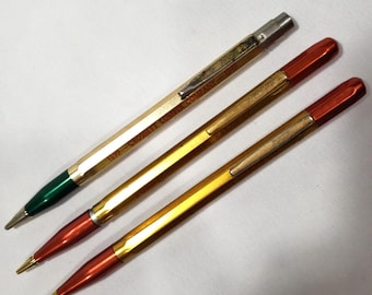 May include: Three mechanical pencils with hexagonal barrels. The top pencil is gold-toned with a green tip and text reading 1899 - CROSSETT LUMBER COMPANY - 1949. The middle and bottom pencils feature gold-toned barrels with metallic red tips and caps.