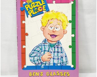 May include: A children's book titled "Ben's Glasses" features a cartoon illustration of a boy with glasses and yellow hair. The book cover has a purple border with the words "The Puzzle Place" and the tagline "Everybody is different in some way."