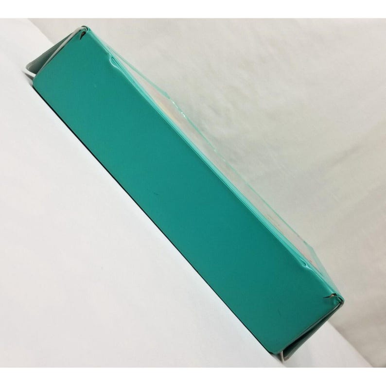 Op de afbeelding: Een rechthoekige, turquoise gekleurde doos met een doorzichtig plastic venster aan de bovenkant. De doos is schuin geplaatst en toont de zijkant en bovenkant. De doos staat op een wit oppervlak.