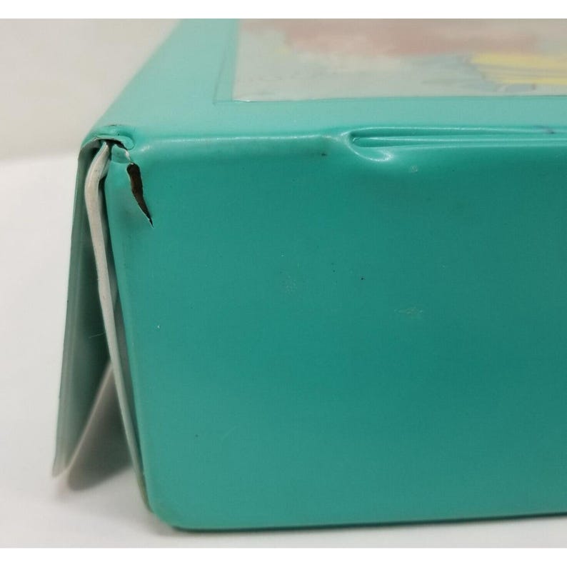 Op de afbeelding: Close-up van een turquoise metalen doos met een witte rand. De doos heeft een deuk en een kleine scheur op de hoek. De doos lijkt leeg te zijn.