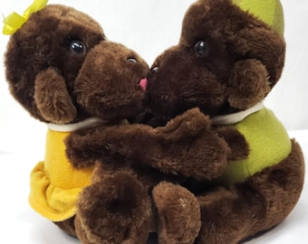 May include: Two brown plush monkeys embracing each other. The left monkey wears a yellow dress with a yellow bow on its head. The right monkey wears a green shirt and a matching green cap. Both have dark eyes and a small pink tongue visible on the left.