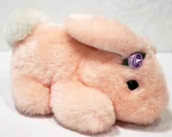 May include: A plush rabbit toy with soft peach-colored fur, a fluffy white tail, and a small purple fabric rose accent attached near the ear. The stuffed animal features a rounded body, long ears, and a single visible black plastic eye.