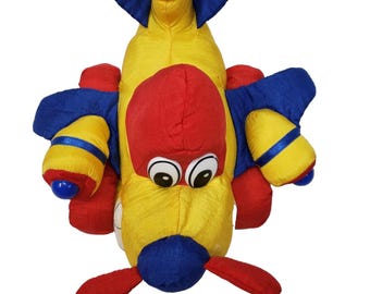 May include: A plush toy airplane in bright primary colors. The toy features a yellow body, red nose and propeller, and blue wings and accents. It has large, cartoonish eyes and is designed to resemble a friendly aircraft.