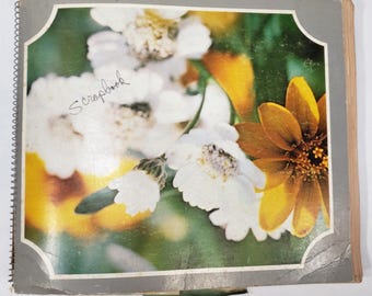 May include: A vintage scrapbook with a floral cover design. The cover features a photograph of white and yellow flowers against a green background. The word "Scrapbook" is handwritten in cursive. The book has a spiral binding.
