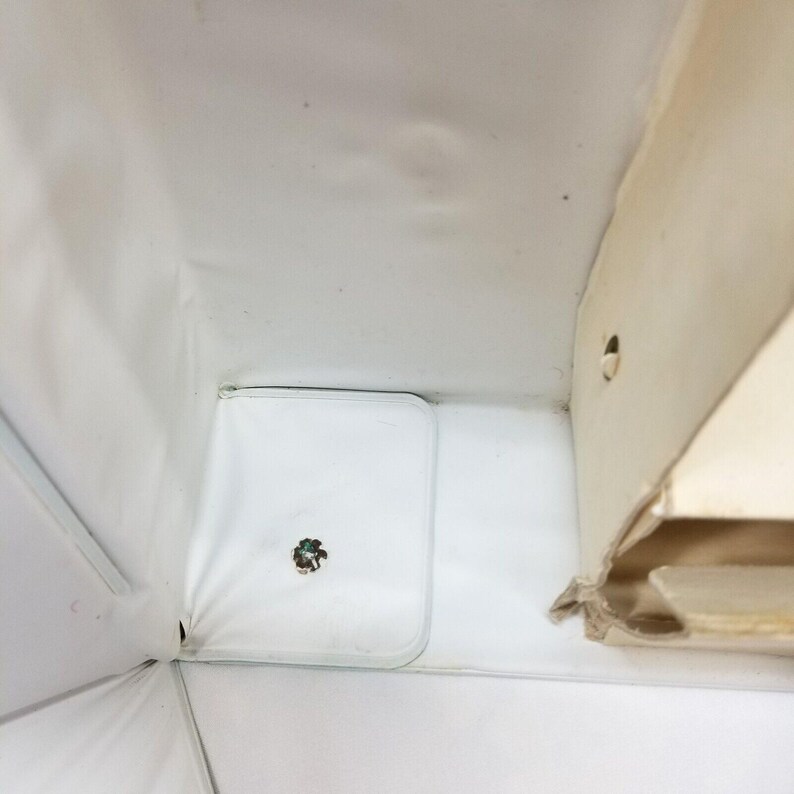 Op de afbeelding: Een bovenaanzicht in een witte doos met een kleine, vierkante, doorzichtige plastic container. De container heeft een klein, donkergekleurd object in het midden. De doos heeft een wit interieur en een beige kartonnen zijkant.