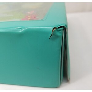 Op de afbeelding: Een turquoise metalen doos met een doorzichtig deksel, met een bloemenontwerp. De doos heeft een deuk en een kleine scheur op de hoek. De doos wordt waarschijnlijk gebruikt voor opslag of weergave.