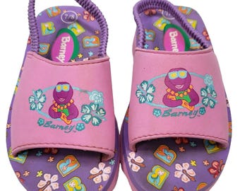 May include: A pair of purple and pink sandals featuring a graphic of the character Barney wearing sunglasses and a lei. The footbed has a repeating pattern of flowers and the letter B. Each sandal includes a pink strap and a purple elastic heel strap.