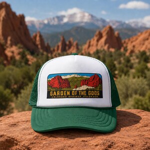 Op de afbeelding: Een witte en donkergroene truckerpet met een rechthoekige grafische patch aan de voorzijde. De patch toont een kleurrijke illustratie van rode rotsformaties en bergen met de tekst GARDEN OF THE GODS COLORADO SPRINGS, COLORADO.