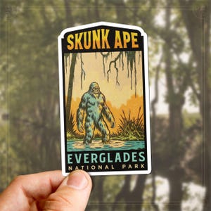 May include: A rectangular sticker featuring a vintage illustration of a Skunk Ape in the Everglades. The text "SKUNK APE" is at the top, and "EVERGLADES NATIONAL PARK" is at the bottom. The Skunk Ape is depicted in a swampy environment.