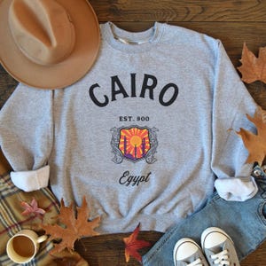 May include: Gray sweatshirt with a black graphic print that says "CAIRO EST. 900 Egypt". The graphic features a shield with a sunset and minarets.