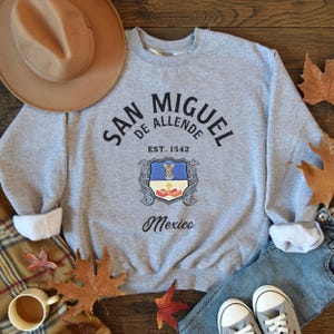 May include: Gray sweatshirt with black text that reads "San Miguel de Allende Est. 1542 Mexico" and a black and white crest with a blue and white flag.