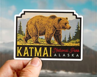 Puede incluir: Una pegatina con una ilustración de un oso pardo caminando por un paisaje con pinos y montañas. La pegatina dice "KATMAI National Park ALASKA" en texto amarillo y rojo sobre un fondo negro.