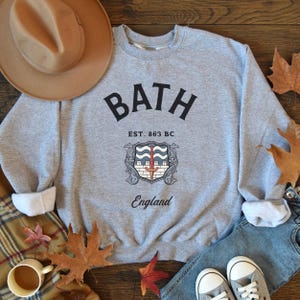 May include: Gray sweatshirt with a black graphic print of the word "BATH" in a large font, with the text "EST. 863 BC" and "England" underneath. The graphic also includes a stylized coat of arms with a sword and waves.