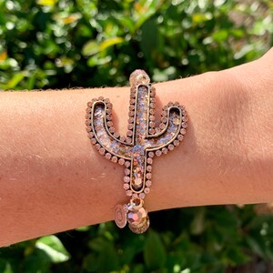 May include: A gold-tone bracelet with a sparkly cactus charm and a dangling charm. The bracelet is made of beads and has a delicate, feminine look.