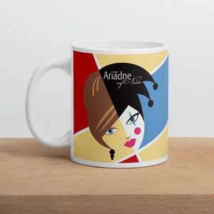 May include: White ceramic mug with a colorful design featuring a woman's face with half of her face painted like a jester. The text "Ariadne auf Naxos" is printed on the mug.