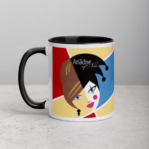 Könnte beinhalten: Weißer Keramikbecher mit schwarzem Griff und Innenraum. Der Becher zeigt eine farbenfrohe Grafik eines stilisierten Gesichts mit braunem Haar, einer Narrenkappe und dem Text "Ariadne auf Naxos". Das Design umfasst rote, gelbe und blaue geometrische Formen.