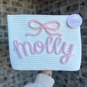May include: White woven storage basket with a pink knitted bow and the name "molly" spelled out in pink yarn.