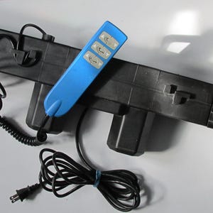 May include: A black, rectangular device with a blue remote control. The remote has three buttons and is connected to the device by a black cord. The device has a power cord and is likely an electronic component.