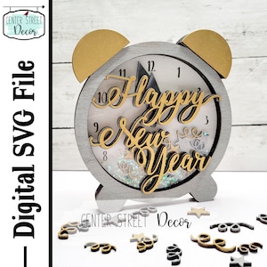 Peut inclure: Une décoration en forme d'horloge grise et dorée pour le Nouvel An. Le cadran de l'horloge porte l'inscription "Happy New Year" en lettres dorées. L'horloge a des accents dorés et est entourée de petites formes décoratives.