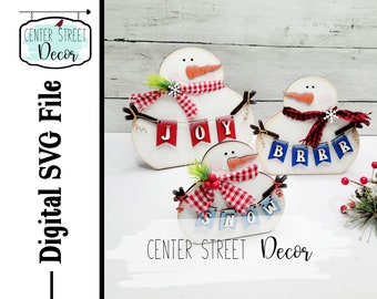 Op de afbeelding: Drie houten sneeuwman ornamenten met rood-wit geblokte sjaals en hoeden. Elke sneeuwman houdt een banner met de woorden "JOY", "BRRR" en "SNOW".