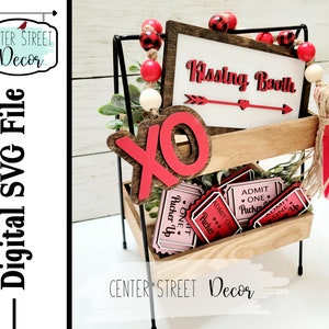 May include: A wooden sign with the text "Kissing Booth" in red letters, with a red arrow and heart. The sign is on a tiered wooden stand with red and white tickets that say "Admit One Pucker Up".