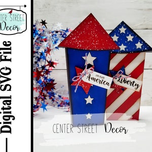 Könnte beinhalten: Zwei hölzerne, rot-weiß-blau bemalte Feuerwerkskörper in Form von Schachteln mit Sternen und den Worten "God Bless America" und "Liberty" darauf.