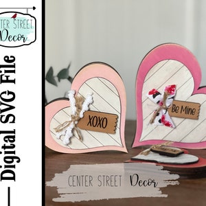 May include: Two pink and white wooden heart-shaped signs with a striped pattern. The sign on the left has a brown tag with the text "XOXO" and the sign on the right has a brown tag with the text "Be Mine".