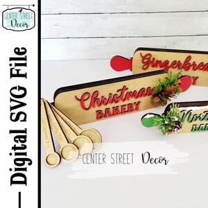May include: Wooden Christmas bakery signs with red and green accents. The signs read "Christmas Bakery" and "Gingerbread". There are also five wooden measuring spoons with the measurements engraved on them.