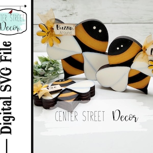 Puede incluir: Dos adornos de madera en forma de abeja con rayas negras y amarillas. La abeja más grande tiene un ala blanca en forma de corazón y la abeja más pequeña tiene un ala negra en forma de corazón. Ambas abejas tienen una pequeña flor y un letrero con texto. El letrero de la abeja más grande dice "Buzzin' Around" y el letrero de la abeja más pequeña dice "Honey Lover".