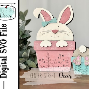 May include: Wooden Easter bunny decor. The bunny is white with pink inner ears and a blue bow. It is in a pink flower pot with floral details. A smaller bunny butt decor is also shown. The text "CENTER STREET Decor" is visible.