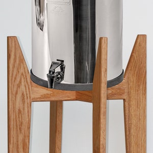 May include: A stainless steel water dispenser with a black faucet sits on a wooden stand with four legs. The stand is made of light brown wood.