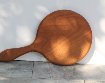 Puede incluir: Un espejo de mano redondo de madera con un mango largo y curvo. El espejo está hecho de madera marrón claro y tiene un acabado liso y pulido.