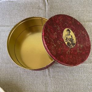 May include: An open, round, vintage tin with a gold interior and a maroon lid. The lid features a marble-like pattern and a small oval image of a person holding a plate. The tin is on a gray patterned surface.