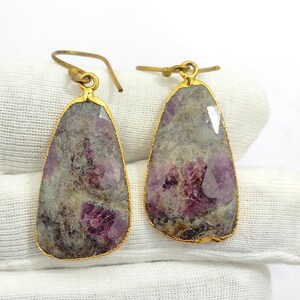 Pode incluir: Um par de brincos em forma de gota. Cada brinco apresenta uma pedra preciosa bruta, cinza e roxa, emoldurada por uma armação de metal dourado. Os brincos têm um fecho de gancho.