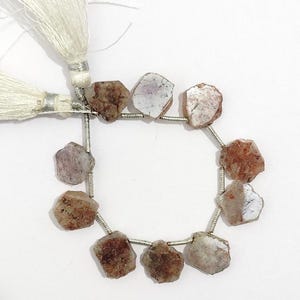 May include: A strand of nine irregular, raw, brownish-pink gemstone slices strung on a silver-coloured wire, featuring two white silk tassels at the top. The stones have a rough, textured surface with visible mineral inclusions and varied shapes.