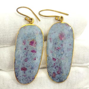 May include: A pair of gold-coloured earrings with large, oval stones. The stones have a light blue-grey base with flecks of deep pink, framed by a thin gold border. The earrings have a simple hook design.