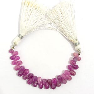 May include: A necklace of polished, oval-shaped, pink gemstones. The gemstones are strung together, forming a curved line. The necklace is attached to two white tassel accents with silver-coloured metal findings. The gemstones vary in shade from light to deep pink.