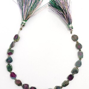 May include: A necklace featuring raw, multi-colored tourmaline beads in shades of green, pink, and gray. The necklace is strung with small silver-colored beads and finished with two decorative tassels with green, purple, and white threads.