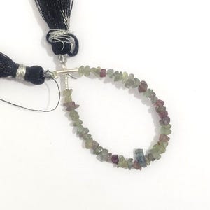 May include: A strand of small, irregular, multi-colored gemstone chips in shades of pale green, olive, and muted purple. The chips are strung on a thin wire, connected to two black fabric tassels with silver-colored metal accents at the top.