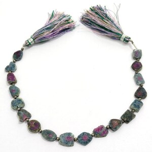 May include: A necklace featuring rough-cut gemstones in shades of blue and magenta. The stones are strung together with small silver beads. The necklace is finished with two tassels in shades of green, purple, and white.