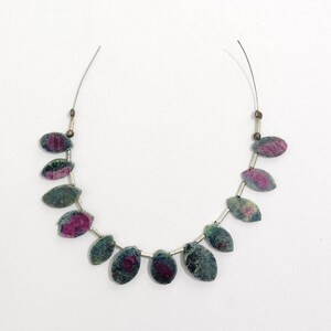 May include: A necklace with oval-shaped beads in shades of green, grey, and magenta. The beads are strung on a thin, silver-coloured wire. The beads have a dark green and grey base with vibrant magenta and pink accents.