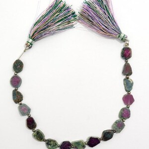 May include: A necklace featuring rough-cut gemstones in shades of green, pink, and purple. The stones are strung together with small silver beads. The necklace is finished with two decorative tassels in green, purple, and white.